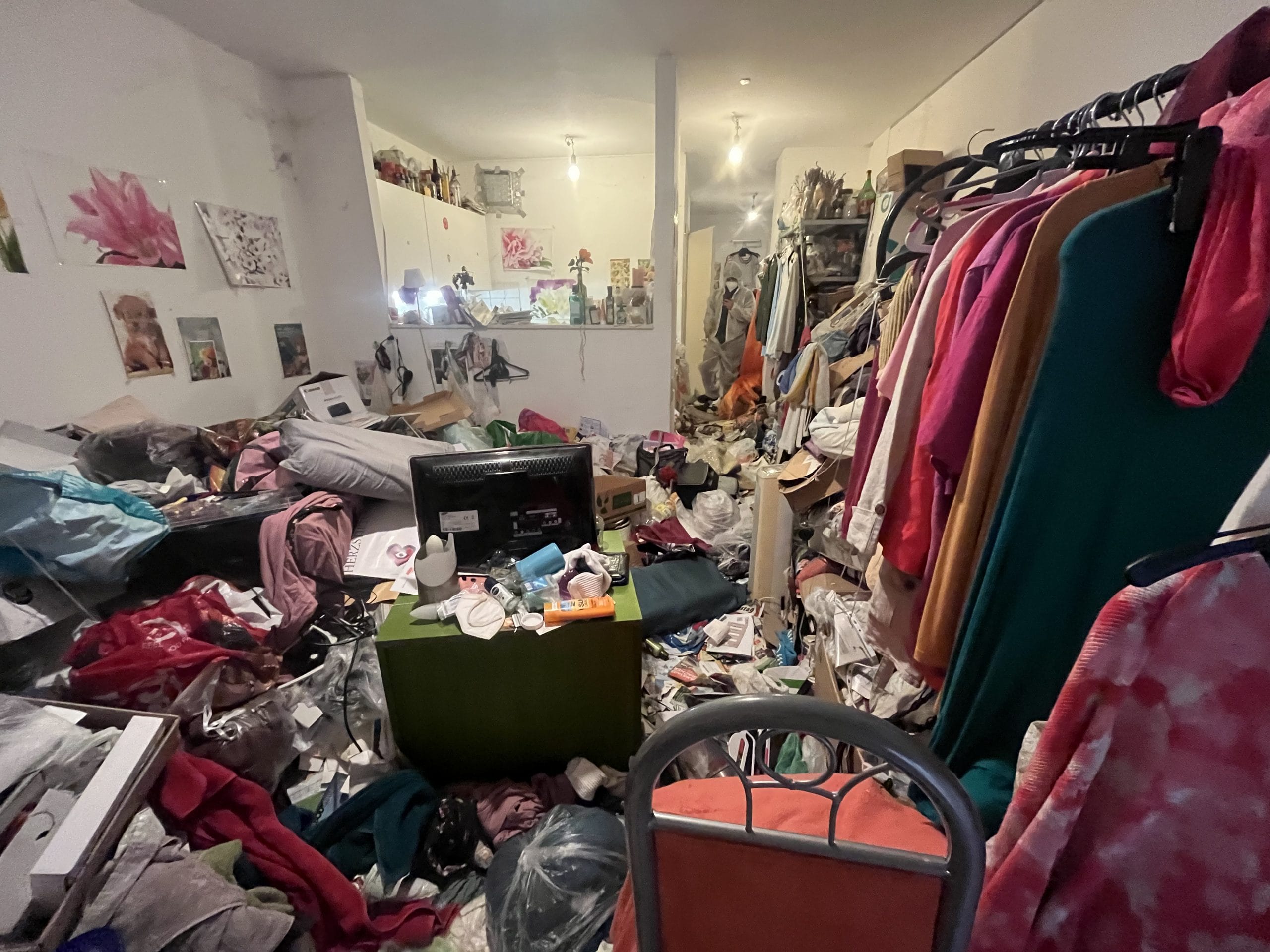 Unordentliches Zimmer, bedeckt mit Kleidung, Müll und verstreuten Gegenständen, auch als 'Messi-Haus' bekannt. Im Hintergrund sind einige Bilder an der Wand zu sehen. Unordentliches Zimmer, bedeckt mit Kleidung, Müll und verstreuten Gegenständen, auch als 'Messi-Haus' bekannt. Im Hintergrund sind einige Bilder an der Wand zu sehen.