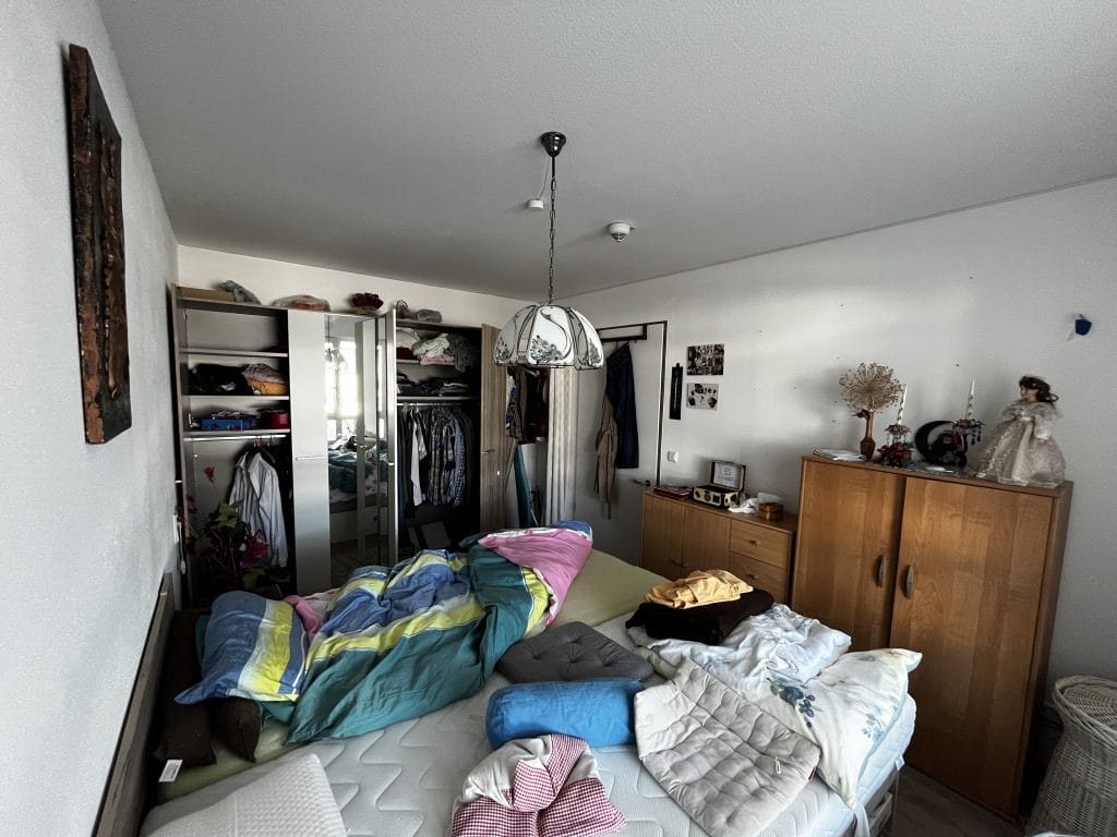 Unaufgeräumtes Schlafzimmer mit einem Kleiderschrank und Gegenständen auf dem Bett, vor Entrümpelung.