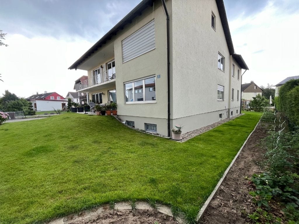 Frisch verlegter grüner Rasen vor einem Mehrfamilienhaus, sauber gepflegter Garten mit Blumentöpfen auf der Terrasse. Frisch verlegter grüner Rasen vor einem Mehrfamilienhaus, sauber gepflegter Garten mit Blumentöpfen auf der Terrasse.