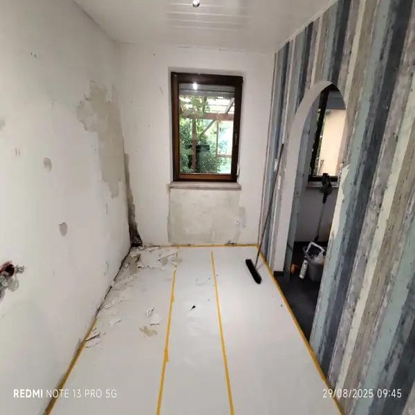 Raum in Renovierung mit entfernten Tapeten, beschädigten Wandflächen und abgedecktem Boden, bereit für weitere Renovierungsarbeiten.